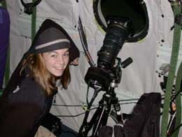A young girl smiles as she stands near what appears to be a long-lens camera on a tripod that is directed out a porthole window.