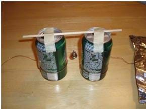 Photo shows two soda cans, placed two inches apart, and connected by a wooden dowel with a bell hanging by a thread from the middle of the dowel.