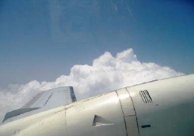 Photo from the window of an airplane shows airplane wing, blue sky and a patch of clouds.