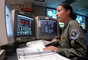 A young woman at a desk with three computer monitors (a space system operator) runs through a checklist during GPS satellite operations.