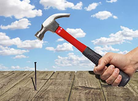 Photograph of a person using a hammer on a nail with a cloudy blue sky background.