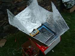 Photo shows a solar oven composed of a box with a solar panel on top, surrounded by angled foil-covered reflective panels.