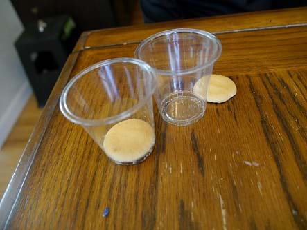 Two clear plastic cups on a wooden table: one cup with a Nilla Wafer sitting next to it; one cup with a Nilla Wafer at the bottom of the cup.