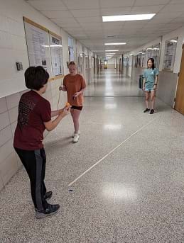 Three students stand in a hallway. One student holds a ping pong ball in his hand, facing away from camera, ready to flick the ball along a path of meter sticks on the ground toward a target identified on the floor with masking tape. A second student stands behind the target observing. A third student stands off to the side, also observing.