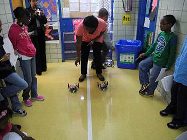 Photo shows teacher releasing two robotic racers as students line a linear racetrack in a school hallway.