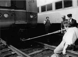 A black and white photo shows a man leaning back on the tracks as he bites on a rope connected to a train car.