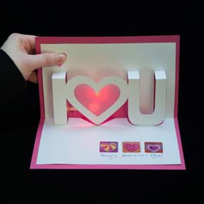 A photograph shows a hand opening a red and white pop-up and light-up valentine card that says “I <heart> you.”  