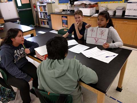 A photograph shows four youngsters sitting around a table. One girl holds a small whiteboard to show the other three kids three different wind turbine sketches.