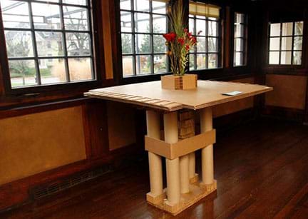 A photograph shows a square table with center support made of corrugated and smooth cardboard in the Prairie style with simple, geometric details that sits in a corner of a room surrounded by a bank of windows with small square panes.