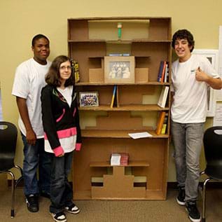 A photograph shows three teens standing next to a six-shelf bookcase that is taller than they are, which they designed and built of brown cardboard.