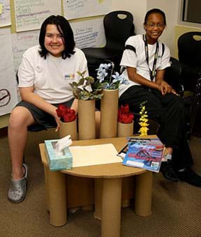 A photograph shows two girls sitting behind a low table that they designed and built of brown cardboard, including upright tubes holding flowers.