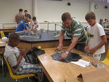 Image of a teacher standing at a counter in a science lab with two young students helping them create parachutes out of black plastic bags and string. The lab table is has supplies on it such as clipboards, paper, pens, tape and safety goggles.  