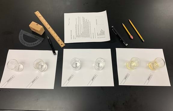 Materials for the activity are laid out on the table. There are three paper placemats with two glasses sitting on each one. The activity worksheet, a protractor, ruler, laser, and pencils are also on the table. 