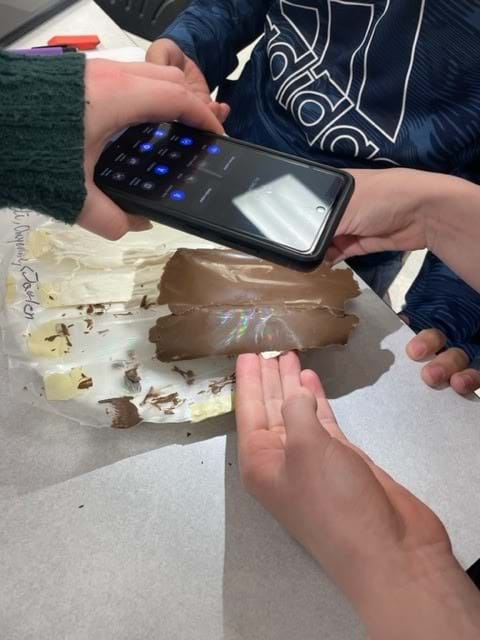 Students shine the flashlight on their phone at the chocolate. A rainbow hologram is visible on the chocolate. 