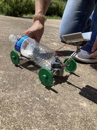A constructed solar rover. A recycled water bottle sits on a frame with four wheels in the sun. A small solar panel attached to the back of the rover extends above the rover.