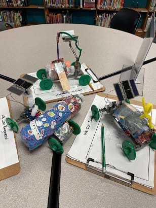 Three solar rover designs made of recycled materials on top of frames with four wheels sit on the table. Each has a small solar panel attached to the back that extends above the rover. The recycled materials for each include a tape- and sticker-covered water bottle, a small plastic bottle with popsicle sticks taped to it, and a tape-covered water bottle with a pipe cleaner attached to it. 