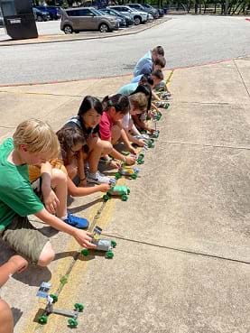 Students are in a line kneeling on the ground next to a line on the sidewalk. They are holding their solar rovers in place at the start line for the race.
