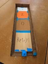 Student prototype with a board modified with four different kinds of surfaces (wood, sandpaper, foam, paper), with different scoring opportunities. A launcher is placed at the front of the board that the player can use to launch the puck. The launcher uses a rubber band slingshot mechanism.