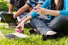 A photograph shows three teenagers sitting on the grass using laptops and cell phones and listening to music with ear buds.