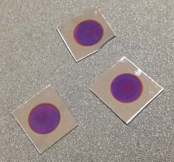 A photograph shows three squares of rigid silver material (sample Bragg mirrors) lying on a gray countertop. Each square has purplish circles etched into its surface.