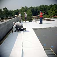 Photo shows four men adhering long white rolls of material to a flat black rooftop.