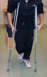 Photo shows a boy using a pair of crutches. Attached to the outer side of one crutch below the hand support is a container—a flat wooden box with the top side open; it contains a notebook.