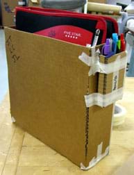 Photo shows an open-topped cardboard box with drafting tape at corner seams; it holds a zipped school notebook/binder. A smaller cardboard box holding three pens is taped to one end of the bigger box.