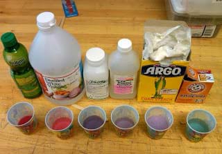 Photo shows a line-up of six small paper cups containing small amounts of red, blue purple and green liquids, next to original containers of test item: lemon juice, white vinegar, isopropyl alcohol, corn starch, baking soda.