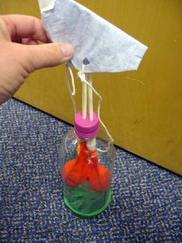 Photo shows a hand placing a folded cloth over two straws sticking out of the top of a two-liter bottle lung model. Photo shows a hand placing a folded cloth over two straws sticking out of the top of a two-liter bottle lung model.
