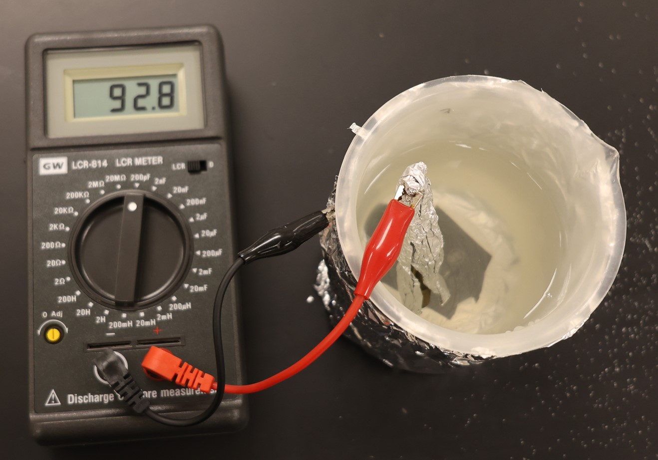 An LCR meter has a black and a red clip attached to it. These clips are attached to the two aluminum sheets used in the experiment. The black clip is hooked to the sheet that is wrapped around the cylinder, while the red clip is attached to the foil that is in the saltwater solution.