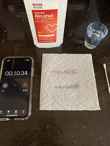 A cell phone timer, paper towel with two wet streaks, a Q-tip, a small glass of water, and a container of rubbing alcohol.