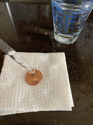 A paper towel with a penny on top of it and two drops of water on the penny that have come from a dropper above. There is a small glass of water in the top right of the image.