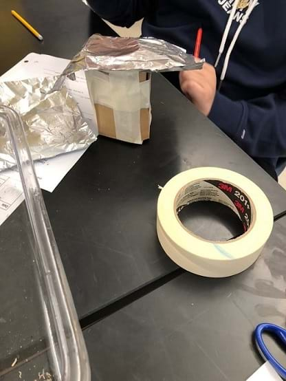 An insulated cup design sits on the table with a roll of tape next to it. The cup is covered by taped-on cardboard, and a piece of foil sits on the top of the cup.