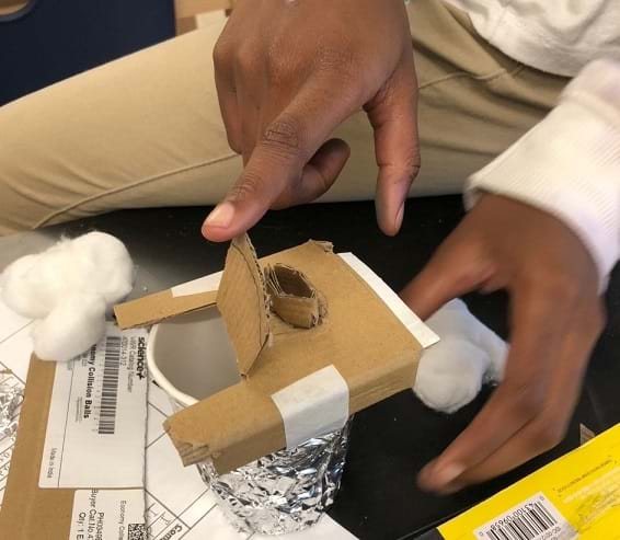 An insulated cup design sits on the table. The cup is covered in foil and has a cardboard lid with a flap that lifts up. A student holds the flap open to show the inside of the cup. 