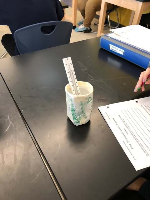 An insulated cup design filled with water sits on the table. The cup is covered in plastic. A thermometer sits inside the cup.