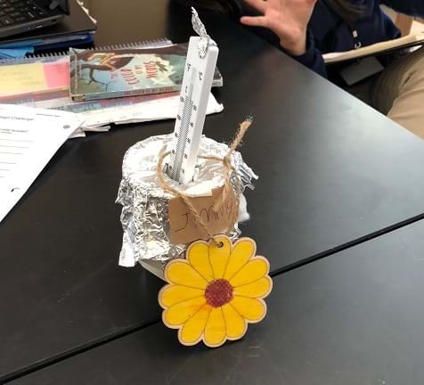 An insulated cup design filled with water sits on the table. The cup is covered in foil, including a foil lid, and is labeled “Jimmy” and has a sunflower ornament next to it. A thermometer breaches the foil lid and sits in the cup.