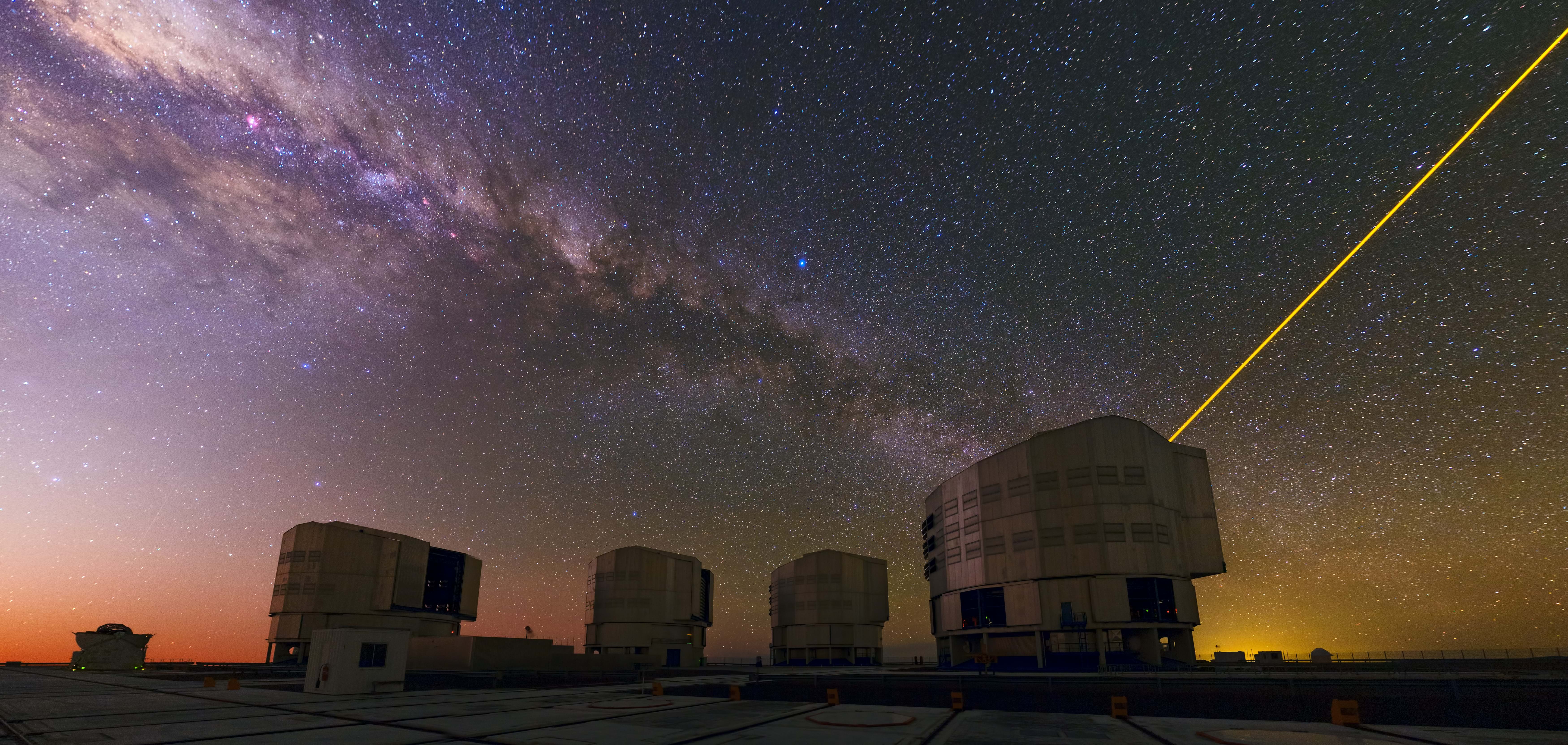 A panorama of ESO’s Very Large Telescope (VLT) atop the platform of Chile’s Cerro Paranal. A mix of colours sweeps across the sky in this image. From the fiery red sunset up to the dusty purple band of the Milky Way.