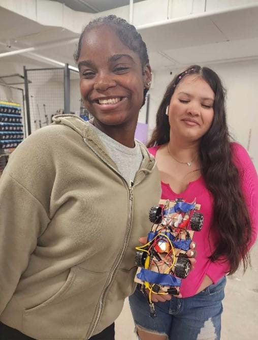A couple of girls standing next to each other with one girl holding the model EV car they designed and built.