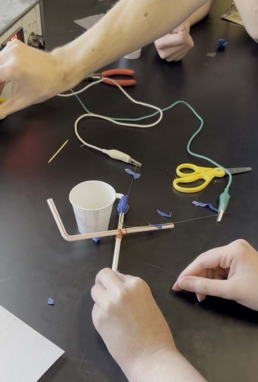 A photo showing wires, tools and other classroom supplies sitting on a lab table with a student-built claw design; the student design is made from straws, tape and nitinol wire; students are holding the claw and controlling a power supply knob to cause the wire to change shape.