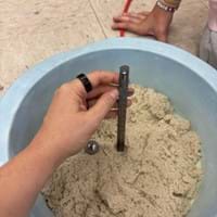 A photo showing a hand holding a metal depth gauge in modeling sand to measure the indentation left by a dropped steel ball. A student’s arm and red cord are visible in the background.