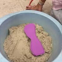 A photo showing a pink foam orthotic insole lying on top of a bin filled with modeling sand. A student’s hand rests beside the container.