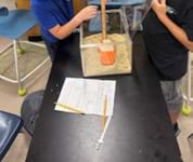 A photo showing two students dropping a fishing weight into a square container of kinetic sand. One student is holding a meter stick next to the container.