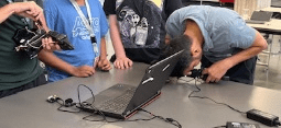 A photo showing a group of students standing around a table in a classroom. One student is leaning over and looking closely at a laptop screen while others observe. Various cables and small electronic components are connected to the laptop.