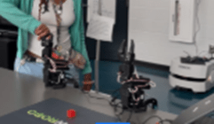A photo showing a student standing at a table in a classroom, adjusting a small robotic arm connected by wires. Another robotic arm sits nearby on the table. A laptop and additional equipment are also visible.