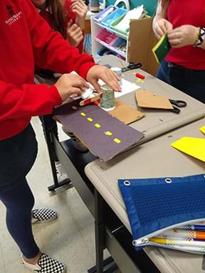 A student gluing a popsicle stick to cup.  The student appears to be building a crossing gate to stop oncoming traffic.