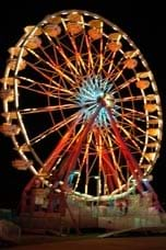 A photograph of a lighted Ferris wheel at night, a rotating upright wheel with passenger cars around the outside of the wheel.