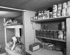 A black and white photo shows two people reading in bunk beds in an underground room with a counter and wall bookcases containing cans and jars of food.