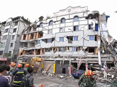 A photograph shows emergency first responders in gear and helmets in front of a six-story residential building (now looks like five-stories) that looks broken, cracked and crumbling.