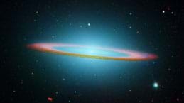 An infrared light image of Sombrero Galaxy. It looks like a large glowing bluish spot surrounded by a pinkish ring and many stars in endless dark space.