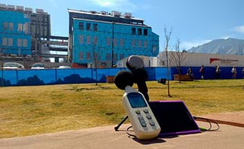 A photograph shows a digital sound level monitor, microphone and digital tablet wired together to measure noise from a construction site seen in the background.